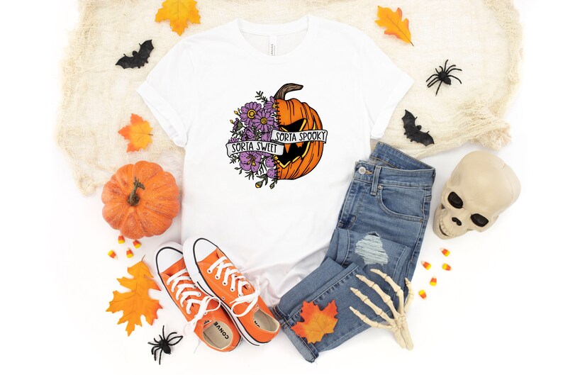 May include: White t-shirt with a graphic of a pumpkin with flowers on one side and a jack-o-lantern face on the other. The text "Sorta Sweet" and "Sorta Spooky" is on a banner around the pumpkin.