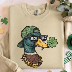 May include: Beige sweatshirt featuring a graphic of a duck wearing sunglasses, a camouflage hat, and a gold chain. The sunglasses read "LUCKY DUCK" and the duck holds a four-leaf clover. The sweatshirt is a Gildan brand.