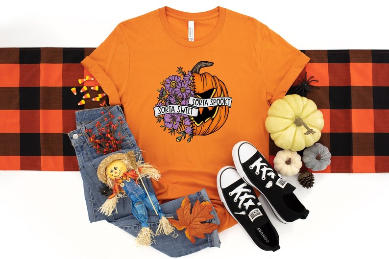 May include: Orange t-shirt with a graphic of a pumpkin with flowers and the text "Sorta Sweet" and "Sorta Spooky". The shirt is laid out on a white background with a plaid blanket, jeans, a scarecrow, and a pair of black sneakers.