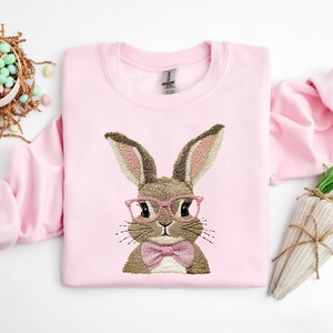 May include: Pink sweatshirt with an embroidered brown bunny wearing pink glasses and a pink bow tie.