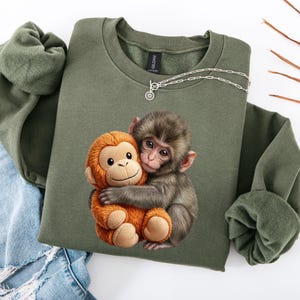 May include: An olive green sweatshirt featuring a graphic of two monkeys hugging. One monkey is brown and the other is orange. A silver necklace with a sun pendant is on the sweatshirt. The sweatshirt is folded on a white surface.