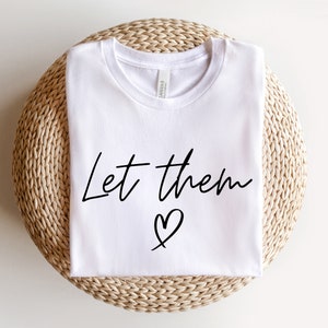 K&ouml;nnte beinhalten: Wei&szlig;es T-Shirt mit dem Text "Let them" und einem Herzsymbol in Schwarz.