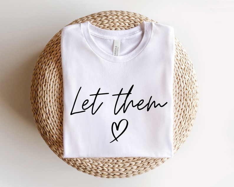 K&ouml;nnte beinhalten: Wei&szlig;es T-Shirt mit dem Text "Let them" und einem Herzsymbol in Schwarz.