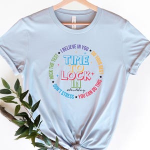 May include: Light blue t-shirt with a circular graphic. The graphic includes the words "TIME TO LOCK IN" in colorful letters, surrounded by phrases like "I BELIEVE IN YOU" and "YOU CAN DO THIS". The hashtag "#testday" is also included.