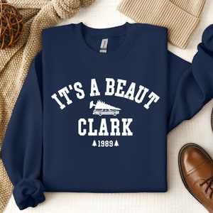 May include: Navy blue sweatshirt with white text that reads "It's a beaut Clark 1989" with a graphic of a car with a Christmas tree on top.