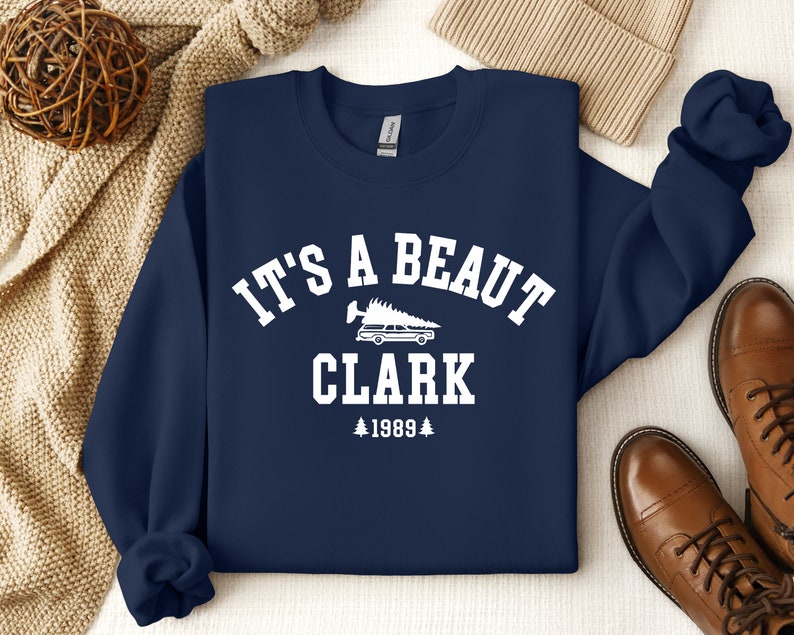 May include: Navy blue sweatshirt with white text that reads "It's a beaut Clark 1989" with a graphic of a car with a Christmas tree on top.