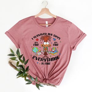 K&ouml;nnte beinhalten: Ein pinkfarbenes T-Shirt mit einer Comicfigur und dem Text "Ich habe 100 Tage &uuml;berlebt. Es ist in Ordnung. Alles ist in Ordnung."