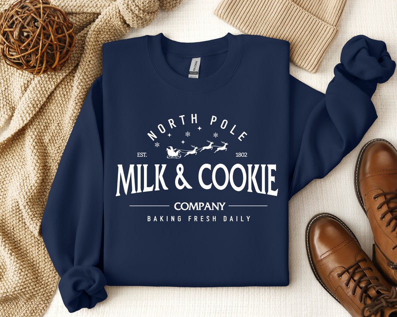 Puede incluir: Sudadera azul marino con texto blanco que dice "North Pole Est. 1802 Milk & Cookie Company Baking Fresh Daily". El texto est&aacute; rodeado de un c&iacute;rculo con copos de nieve y una peque&ntilde;a imagen de un trineo tirado por renos.