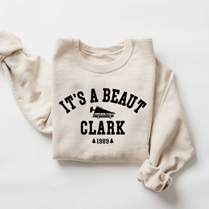 May include: A beige sweatshirt with a black graphic print that says "It's a beaut Clark 1989" with a small image of a car with a Christmas tree on top.