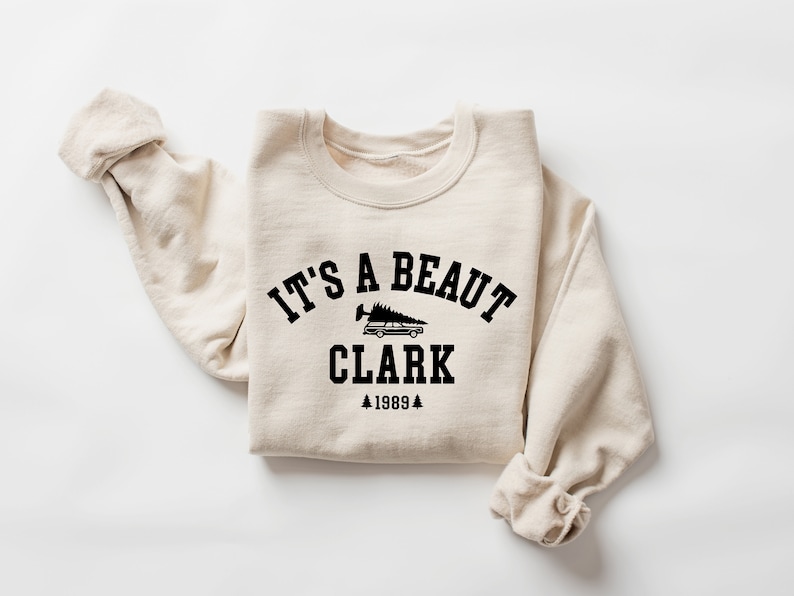 May include: A beige sweatshirt with a black graphic print that says "It's a beaut Clark 1989" with a small image of a car with a Christmas tree on top.