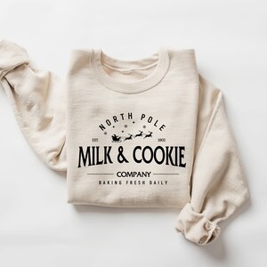 Puede incluir: Una sudadera beige con un dise&ntilde;o gr&aacute;fico negro que dice "North Pole Milk & Cookie Company Baking Fresh Daily" con una ilustraci&oacute;n de Santa Claus y renos.