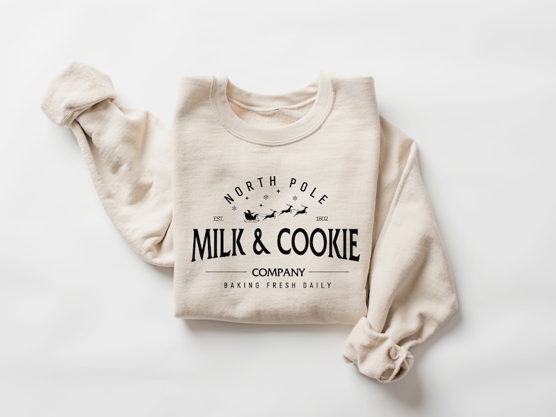 Puede incluir: Una sudadera beige con un dise&ntilde;o gr&aacute;fico negro que dice "North Pole Milk & Cookie Company Baking Fresh Daily" con una ilustraci&oacute;n de Santa Claus y renos.