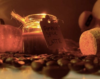 May include: A small glass jar filled with a dark brown liquid, labeled "Pumpkin Spice Latte". The jar is sitting on a bed of coffee beans.