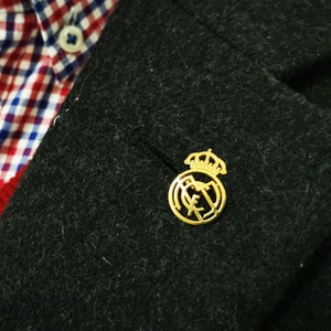 May include: A gold-colored pin with a crown and the letters "R.M." inside a circle. The pin is attached to a black blazer.