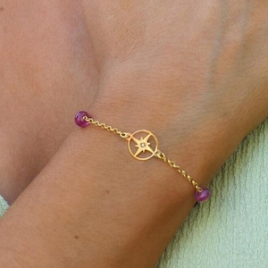 May include: Gold bracelet featuring a compass design and small pink beads. A matching necklace with a compass pendant and blue beads is also visible. The jewellery is displayed on a person's arm and neck.