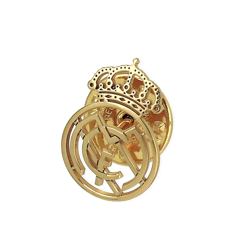 May include: A gold-colored metal pin with a detailed design of a soccer team logo. The logo features a crown and the letters "Real Madrid" in a circular design.