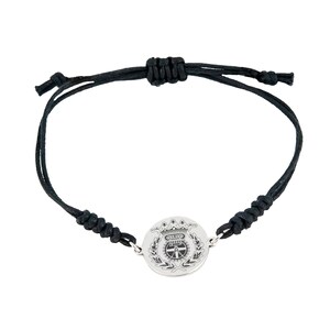 May include: A black cord bracelet with a silver circular charm featuring a detailed engraved design. The charm has a crown, a building, a wheel, and a wreath.