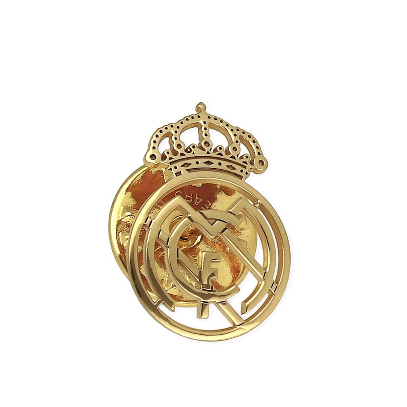 May include: A gold-colored metal pin featuring the Real Madrid crest with a crown on top. The crest is a circular design with a football inside and the letters "F.C.M." around the outside.
