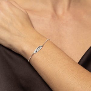 Puede incluir: Un brazalete de cadena de plata con un pequeño dije rectangular. El dije tiene una superficie texturizada y un brillo sutil.
