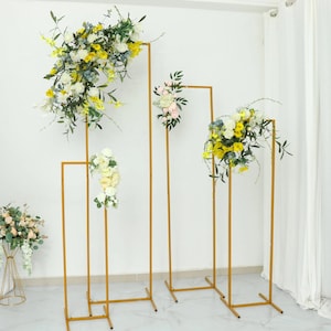 May include: Four gold metal arch stands with different heights, decorated with artificial flowers in shades of yellow, white, and pink. The stands are arranged in a row on a white floor.