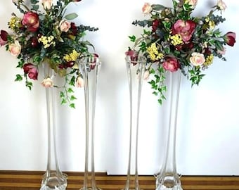 May include: Four tall, clear glass vases with a flared base and a narrow neck. Each vase is filled with a bouquet of artificial flowers in shades of pink, red, and yellow.