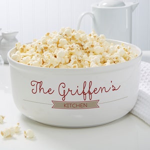 May include: A white ceramic popcorn bowl filled with fluffy popcorn. The bowl has a wide rim and a decorative design in burgundy that reads "The Griffen's KITCHEN". A white teapot and pitcher are in the background.