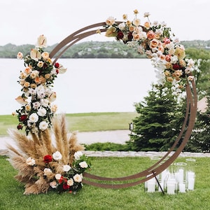 May include: A rustic wooden archway decorated with a floral arrangement of white, pink, and red roses, white orchids, and pampas grass. The archway is set up outdoors against a backdrop of a lake and green grass.