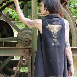 May include: Black sleeveless vest with a gold geometric design on the back. The vest is worn over a patterned top and black lace leggings. The background features a large, rusty metal structure with gears and springs.