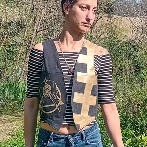 May include: A black and gold vest with a unique design. The vest features a skull and pentagram graphic on one side and a gold cross pattern on the other. The person is wearing a black and white striped shirt and blue jeans.