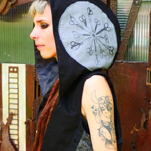 May include: Black hooded vest with a gray circular design on the hood featuring a pattern of scissors. The vest has a dark gray patterned panel on the front. The person in the image has tattoos on their arm and red dreadlocks.