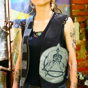 May include: Black vest with a gray skull and triangle design, worn over a black top and paired with black denim shorts. The vest has a patchwork design on the shoulders and a button closure. The person is wearing jewelry and has tattoos on their arms.