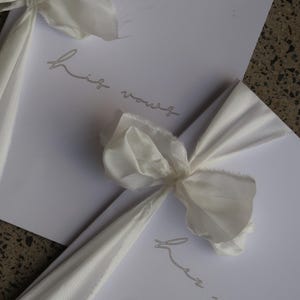 May include: Two white cards with the text "his vows" and "her vows" written in cursive on them. The cards are tied together with a white ribbon.