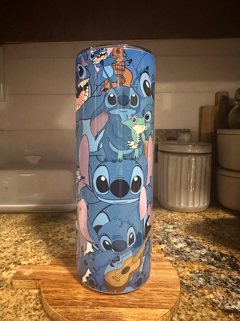 Puede incluir: Un vaso de acero inoxidable azul con un patr&oacute;n repetido de Stitch, el personaje extraterrestre de Lilo & Stitch, en varias poses. Stitch est&aacute; sosteniendo una guitarra, una rana y se muestra en otras poses juguetonas.