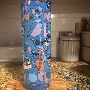 Puede incluir: Un vaso de acero inoxidable azul con un estampado colorido de Stitch de Lilo & Stitch. El estampado presenta a Stitch en varias poses, incluyendo tocando un ukelele.