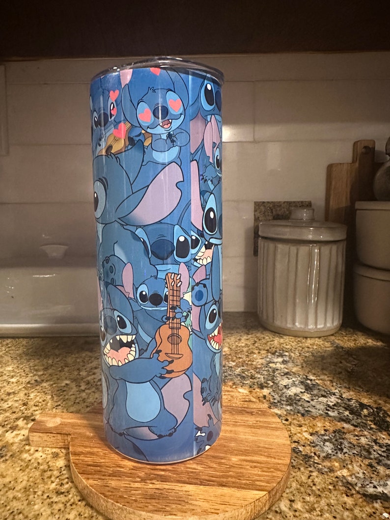 Puede incluir: Un vaso de acero inoxidable azul con un estampado colorido de Stitch de Lilo & Stitch. El estampado presenta a Stitch en varias poses, incluyendo tocando un ukelele.