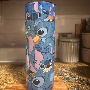 Puede incluir: Un vaso de acero inoxidable azul con un patr&oacute;n colorido de personajes de Stitch de la pel&iacute;cula de Disney Lilo & Stitch. El vaso est&aacute; sobre un posavasos de madera.