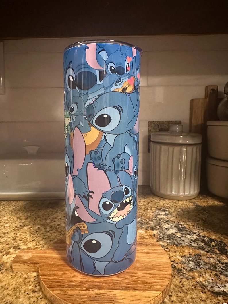 Puede incluir: Un vaso de acero inoxidable azul con un patr&oacute;n colorido de personajes de Stitch de la pel&iacute;cula de Disney Lilo & Stitch. El vaso est&aacute; sobre un posavasos de madera.