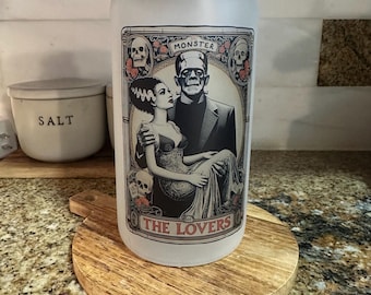 Puede incluir: Un tarro de vidrio esmerilado con tapa de madera. Presenta una ilustración en blanco y negro de Frankenstein y su novia, con las palabras "MONSTER" y "THE LOVERS". Se encuentra sobre un posavasos redondo de madera.