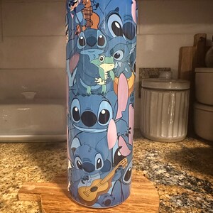 Puede incluir: Un vaso de acero inoxidable azul con un patr&oacute;n repetido de Stitch, el personaje de Lilo & Stitch, tocando el ukelele, sosteniendo una rana y en otras poses.