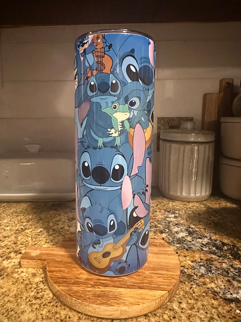 Puede incluir: Un vaso de acero inoxidable azul con un patr&oacute;n repetido de Stitch, el personaje de Lilo & Stitch, tocando el ukelele, sosteniendo una rana y en otras poses.