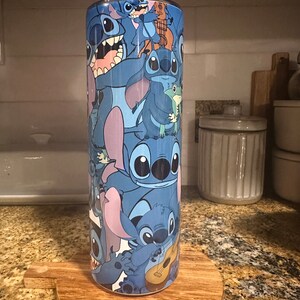 Puede incluir: Un vaso de acero inoxidable azul con un patr&oacute;n colorido de personajes de Stitch de la pel&iacute;cula de Disney Lilo & Stitch. El vaso est&aacute; sobre un posavasos de madera.