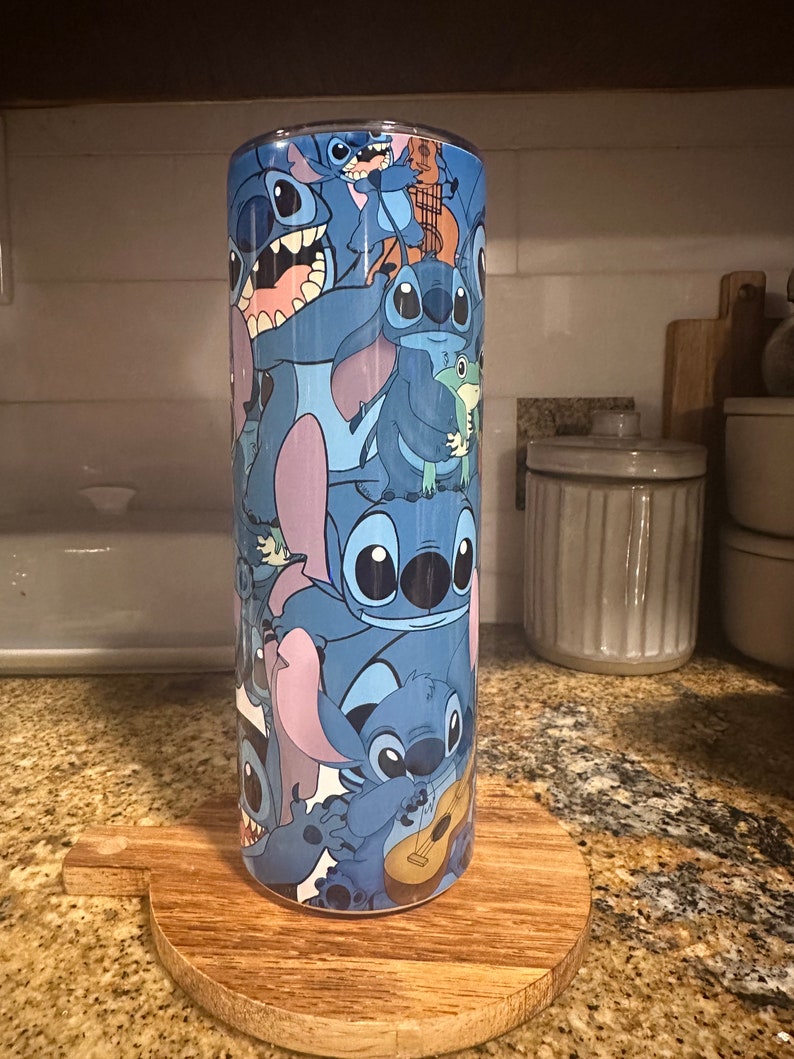 Puede incluir: Un vaso de acero inoxidable azul con un patr&oacute;n colorido de personajes de Stitch de la pel&iacute;cula de Disney Lilo & Stitch. El vaso est&aacute; sobre un posavasos de madera.
