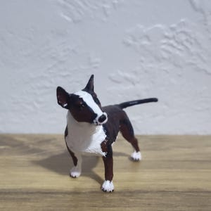 May include: A figurine of a Bull Terrier dog, featuring a brown and white coat. The dog is standing on a wooden surface, with a white wall in the background. The dog's ears are black, and its tail is black.