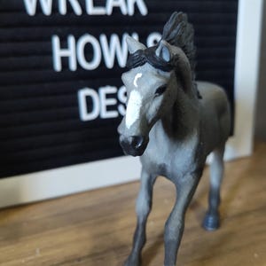 May include: A gray horse figurine with a white blaze on its face and a white marking on one front leg. The horse has a dark gray mane and dark hooves. Behind the figurine is a black felt letter board with the white text WREAK HOWS DES.