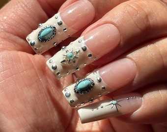 May include: Long square-shaped artificial nails featuring a French manicure base with cream-coloured tips. The tips are decorated with silver studs, turquoise cabochons, and silver star-shaped charms with small turquoise centres.