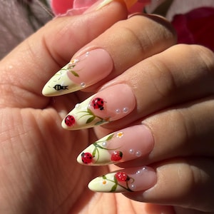 May include: Long, almond-shaped artificial nails with a light yellow base and a French tip design. The nails feature ladybug and floral artwork, with small white pearls. The nails are a unique and artistic accessory.