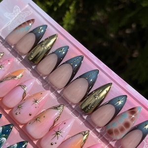 May include: A set of 10 false nails with a variety of designs. The nails are a light pink color with gold accents and a blue ombre effect. Some nails have a star design, while others have a swirl design.