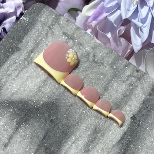 May include: A set of press-on toenails with a pink and yellow design. The big toe nail features a floral embellishment. The nails are arranged on a gray, speckled surface, with a blurred floral background.