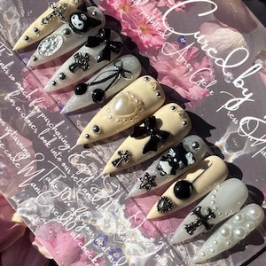 May include: A collection of long, pointed artificial nails with various designs. Some nails are beige, others are white, and some are glittery. Embellishments include black bows, crosses, pearls, and a cartoon character. The background includes text and flowers.