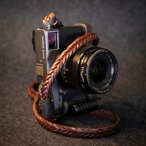 Può includere: Una fotocamera Fujifilm X-Pro2 nera e argento con una tracolla in pelle intrecciata marrone. L'obiettivo della fotocamera è nero con dettagli argentati. La tracolla è avvolta attorno al corpo della fotocamera.